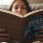 A person reads "Hamlet" by William Shakespeare. The book cover depicts a silhouette of Hamlet holding a skull