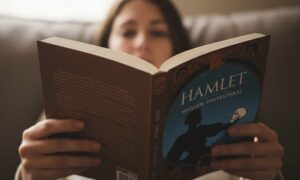 A person reads "Hamlet" by William Shakespeare. The book cover depicts a silhouette of Hamlet holding a skull