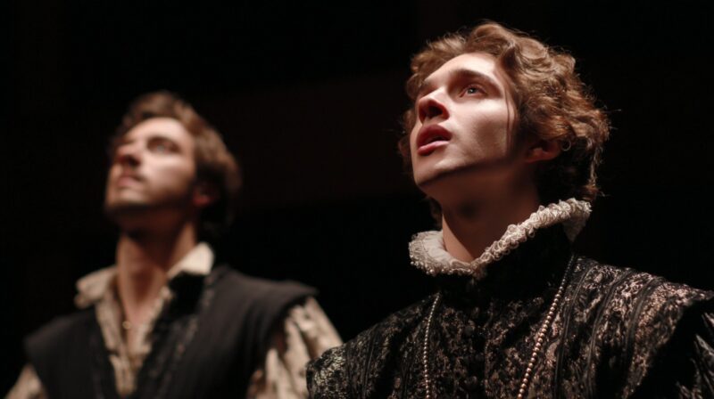 Two actors in Renaissance style costumes perform on a dark stage, lit dramatically from above