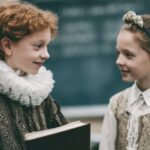 Two children in Shakespearean-style costumes smiling at each other in a classroom, one holding a book, with a chalkboard in the background