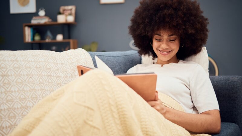 A woman reads a book under a blanket on a sofa at home